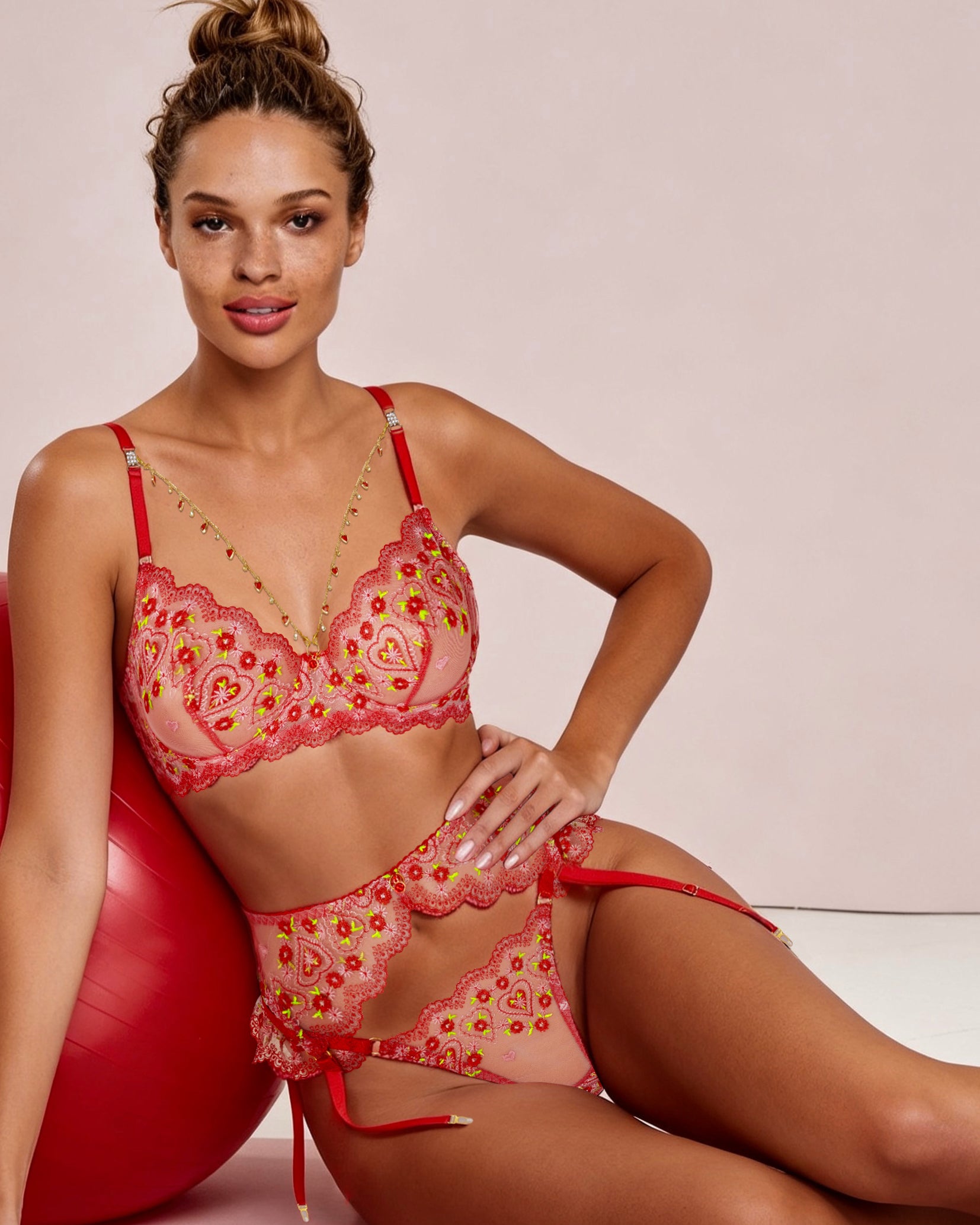 A woman models a two-piece lingerie set consisting of a red and pink paisley-patterned bra and matching g-string bottoms, posed next to a red exercise ball against a light pink background.
