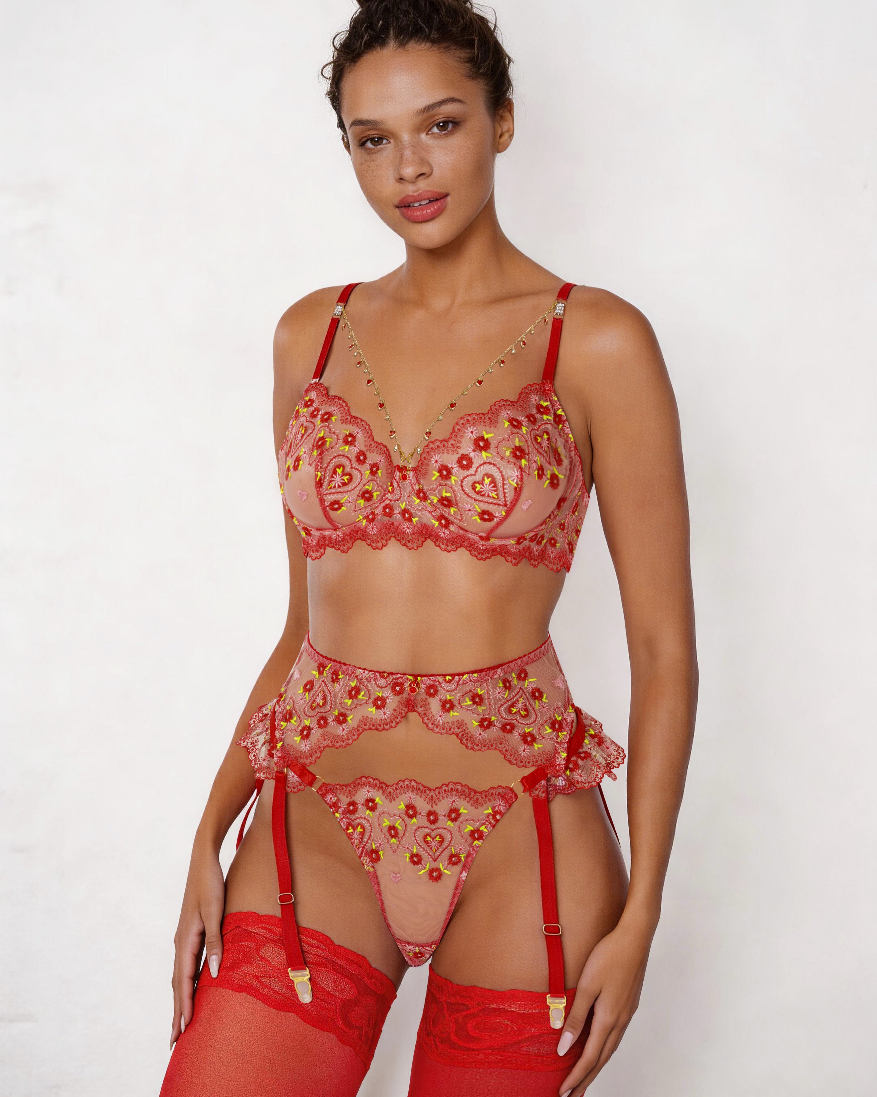 A model wearing a three-piece sheer lace lingerie set in red and pink with gold embroidered details, including a bra, garter belt, thong, and red thigh-high stockings, against a plain white background.