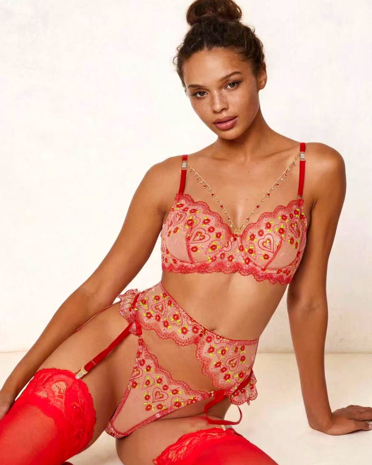 A model wearing a red and pink floral lace lingerie set with matching red thigh-high stockings, posed against a light textured wall.