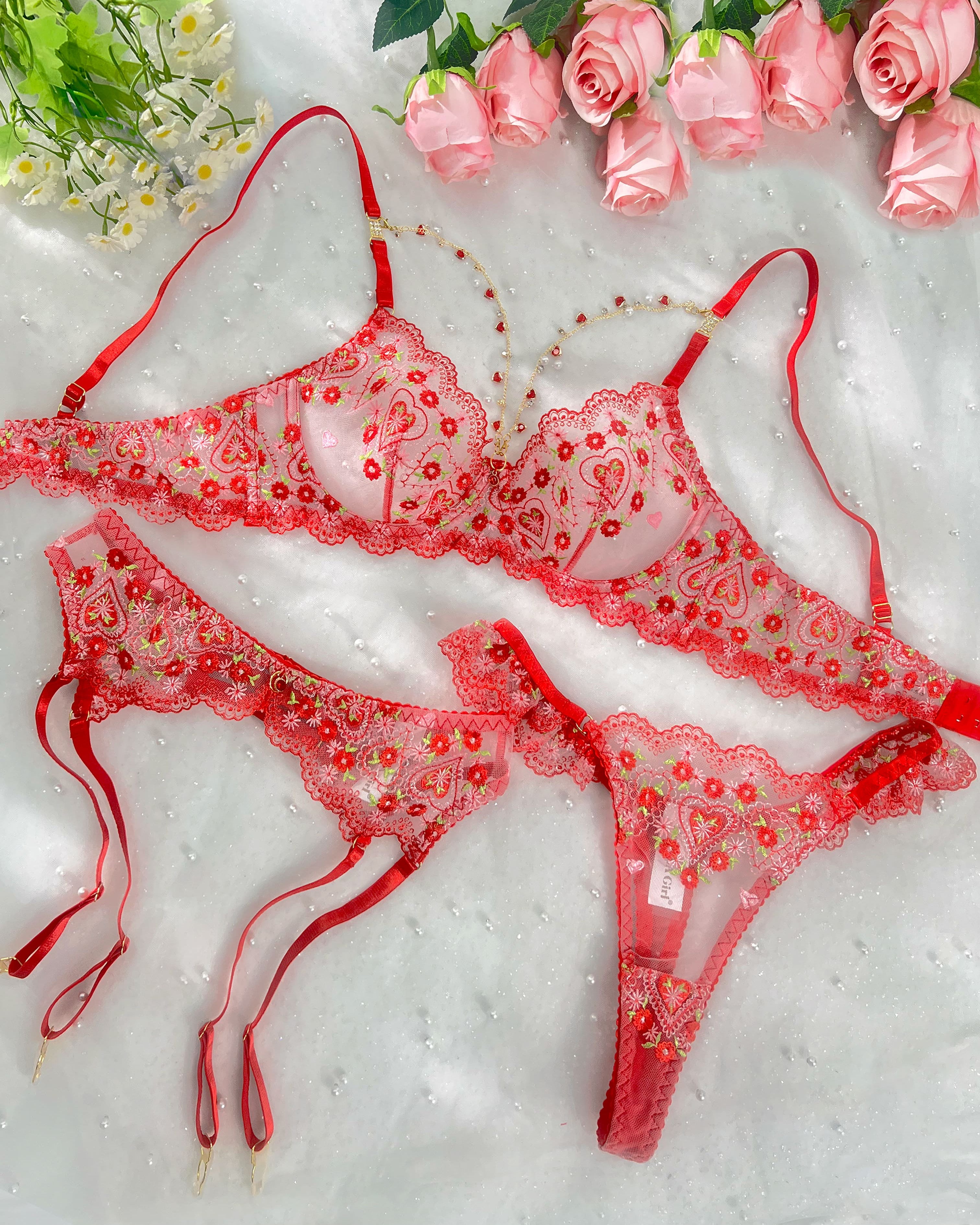 A red sheer lace lingerie set with embroidered floral details and red garter straps, arranged on a white textured fabric background with pink roses and small white flowers as styling props.