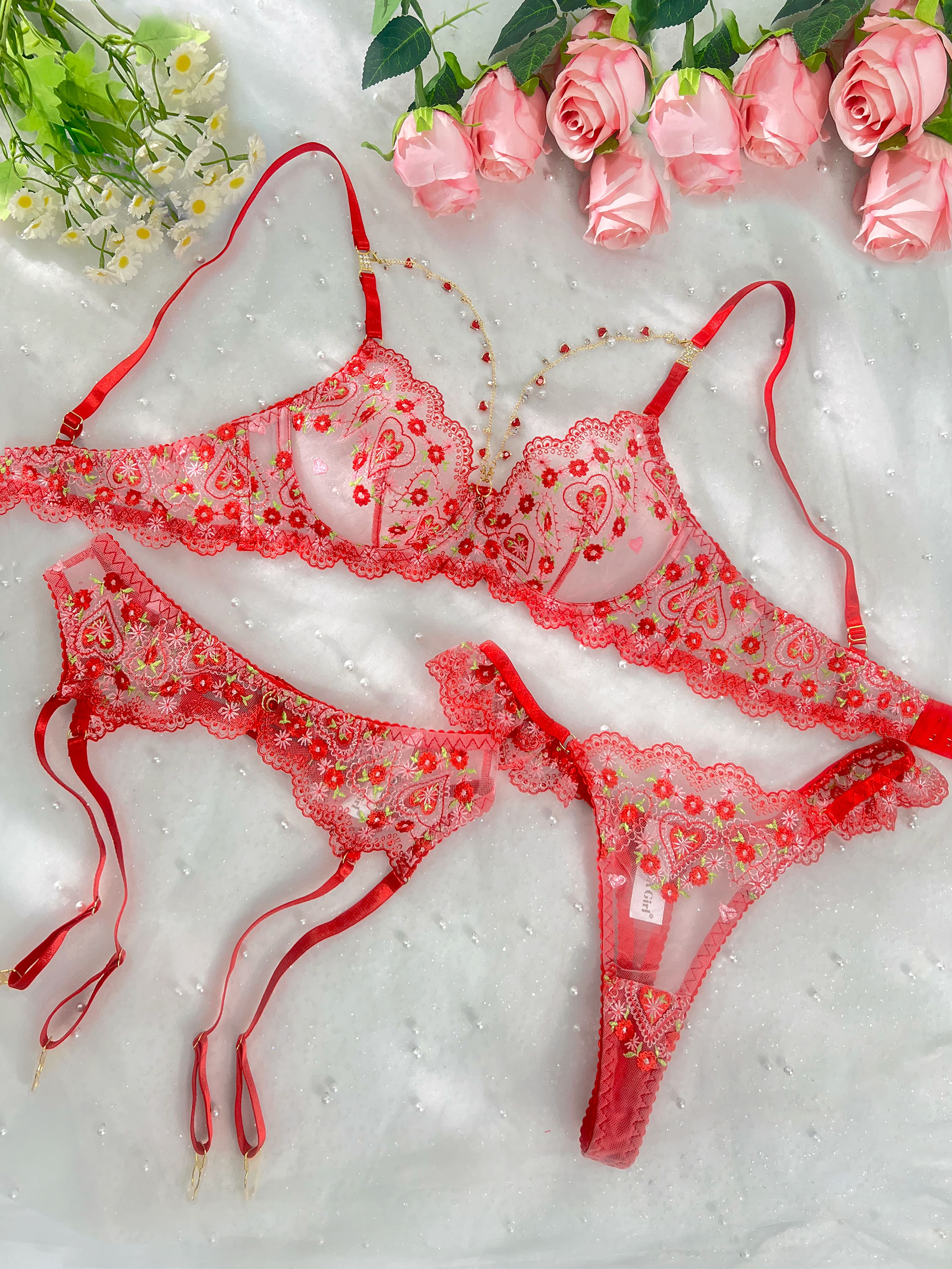 A red sheer lace lingerie set with embroidered floral details and red garter straps, arranged on a white textured fabric background with pink roses and small white flowers as styling props.