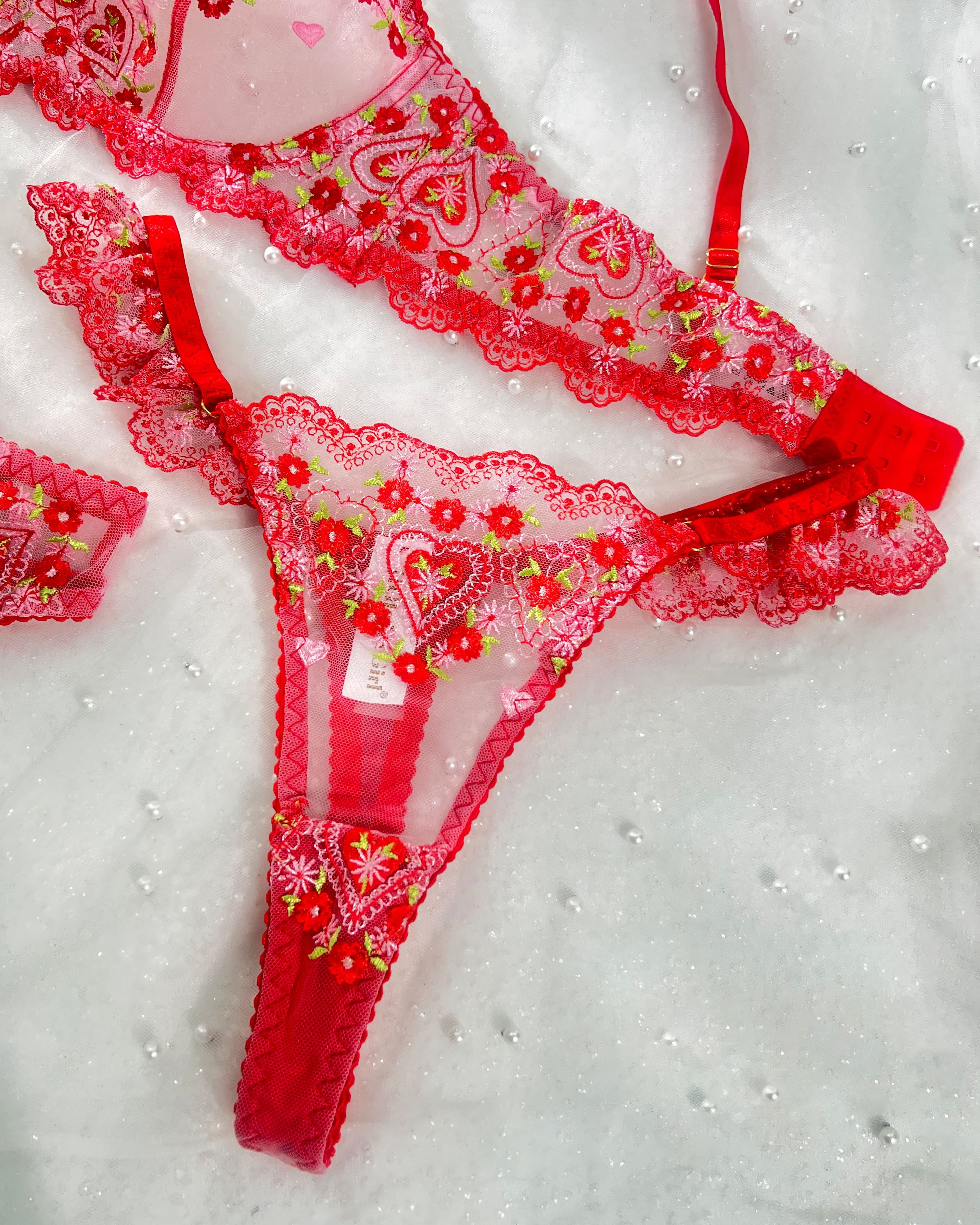 Red lace lingerie set with floral embroidery and heart motifs, laid flat on a white textured background.