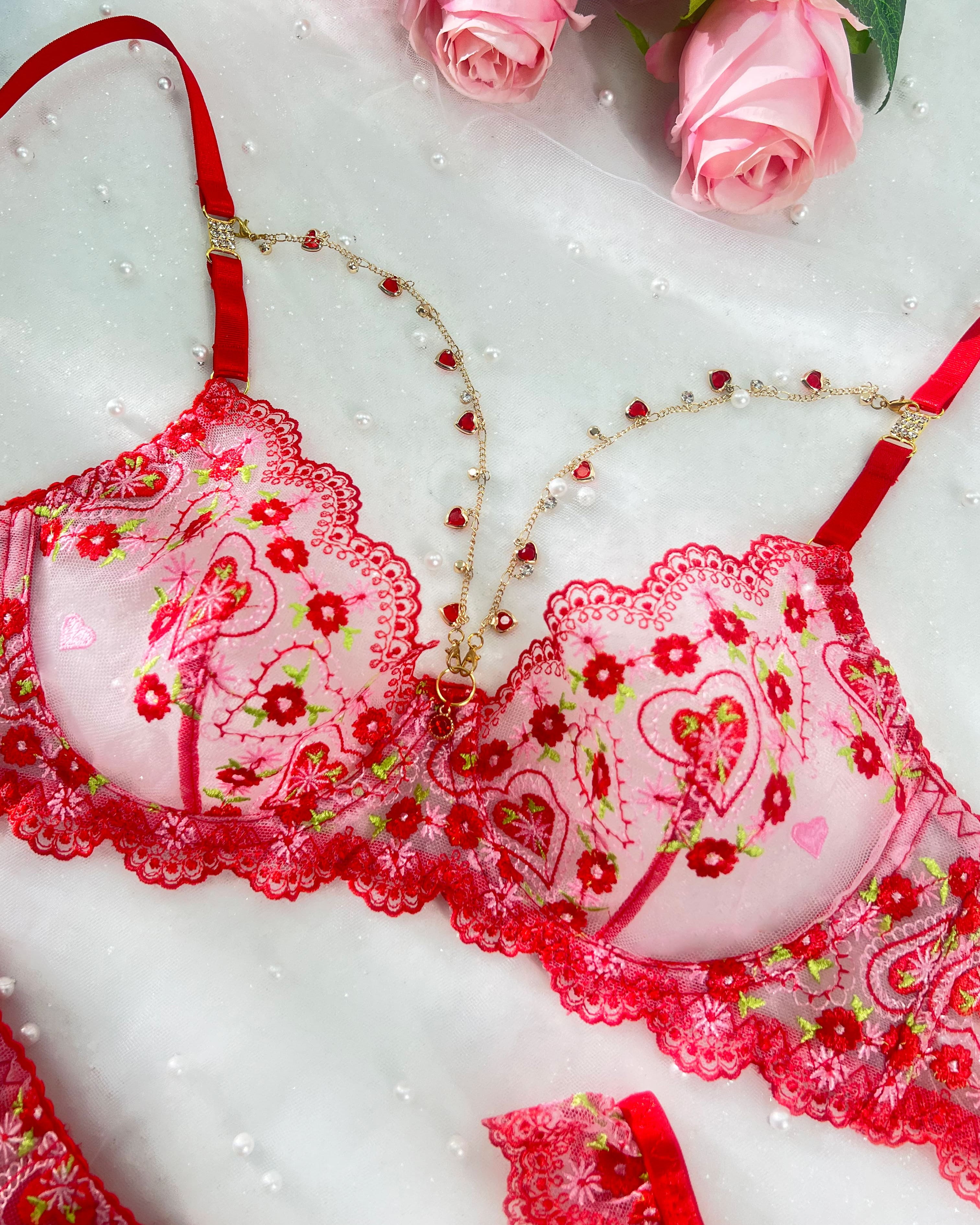 Red and pink lace lingerie set with floral embroidery, heart motifs, and delicate beaded chains, styled with pink roses on a white textured background.