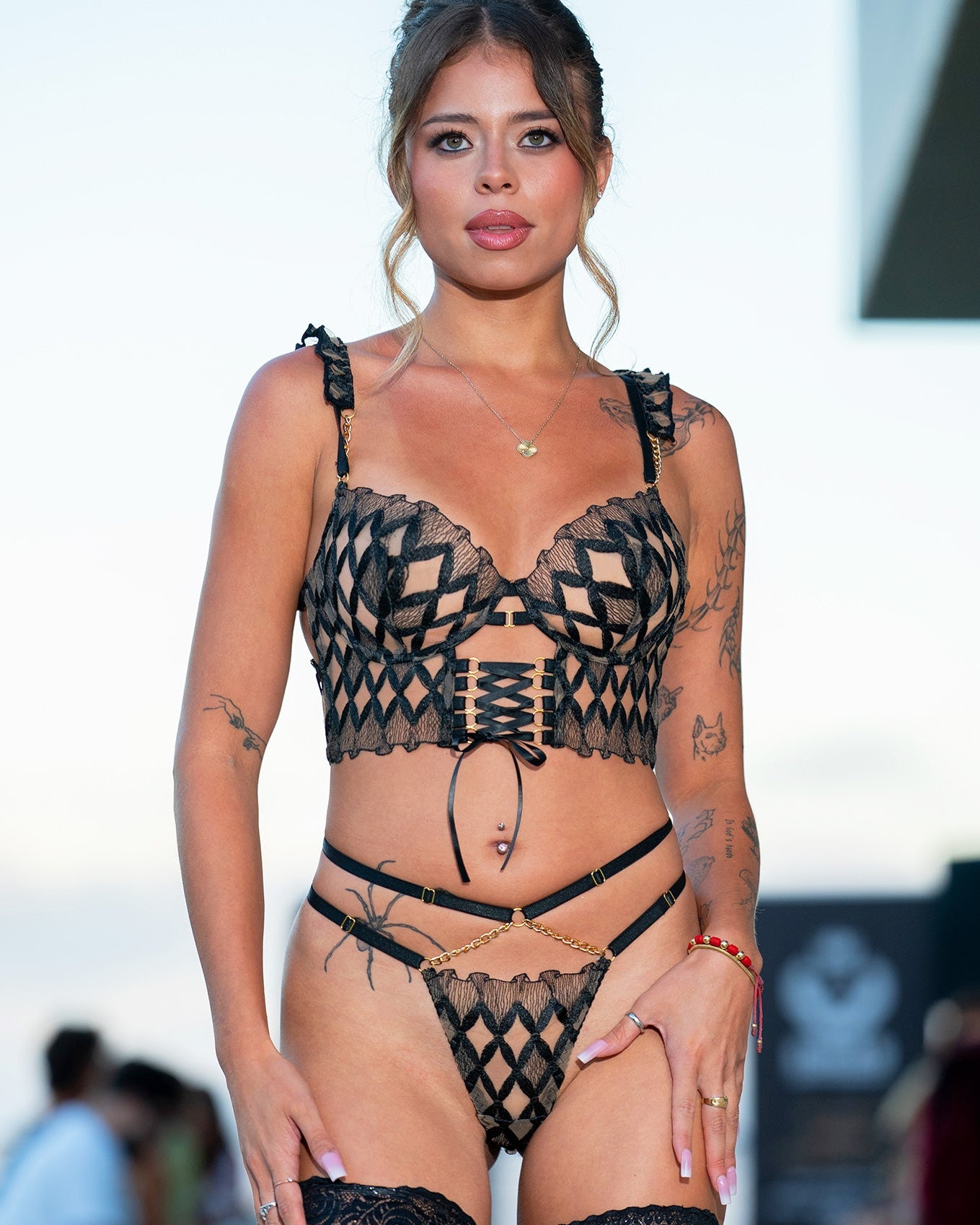 A woman modeling a black lace lingerie set with a geometric pattern, including a bra, thong, garter belt, and thigh-high stockings, set against a blurred outdoor background.