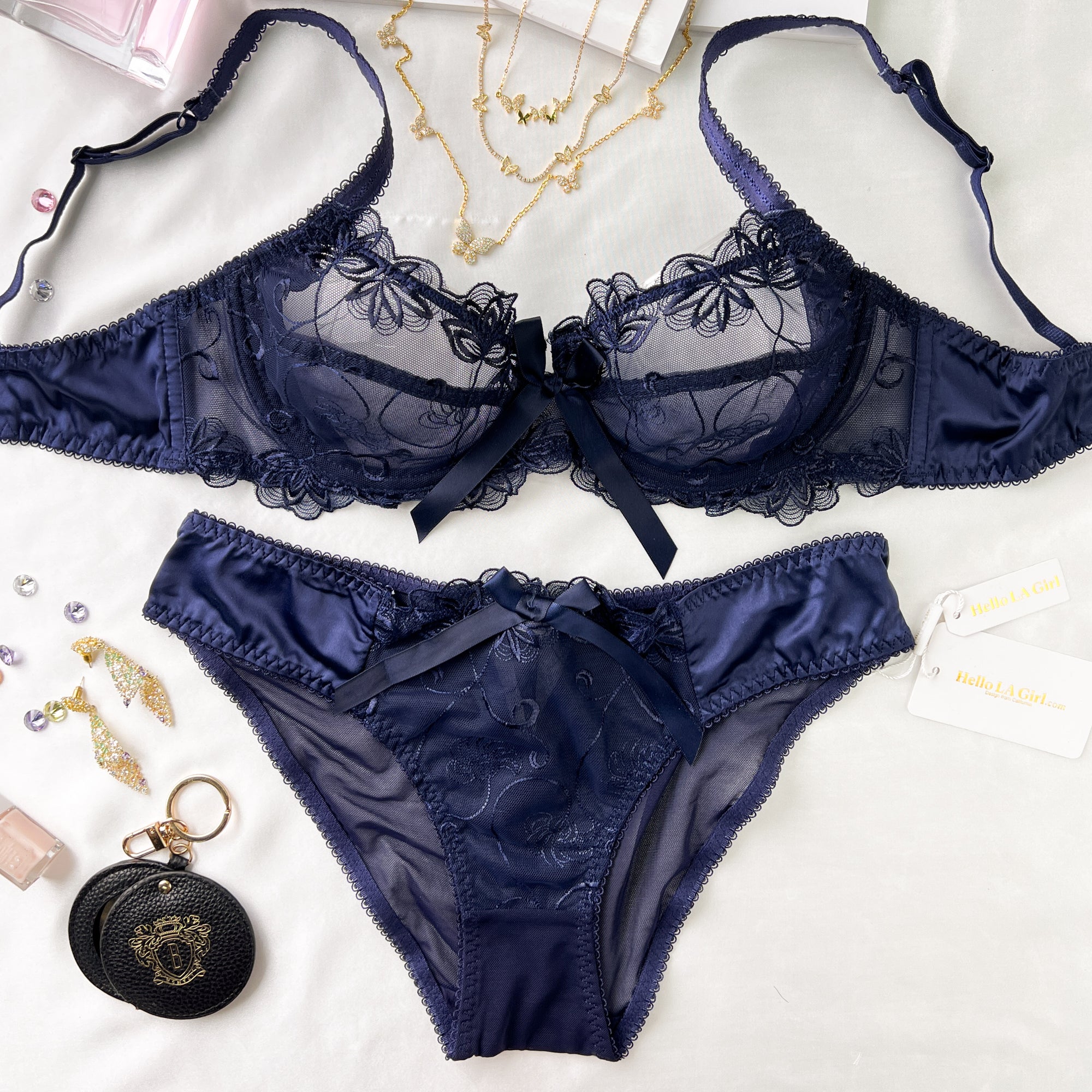 A navy blue lace and satin lingerie set laid flat on a white fabric background, including a bra and matching panties with bow details. Jewelry, a small round black pouch, and a card tag are placed around it as styling props.
