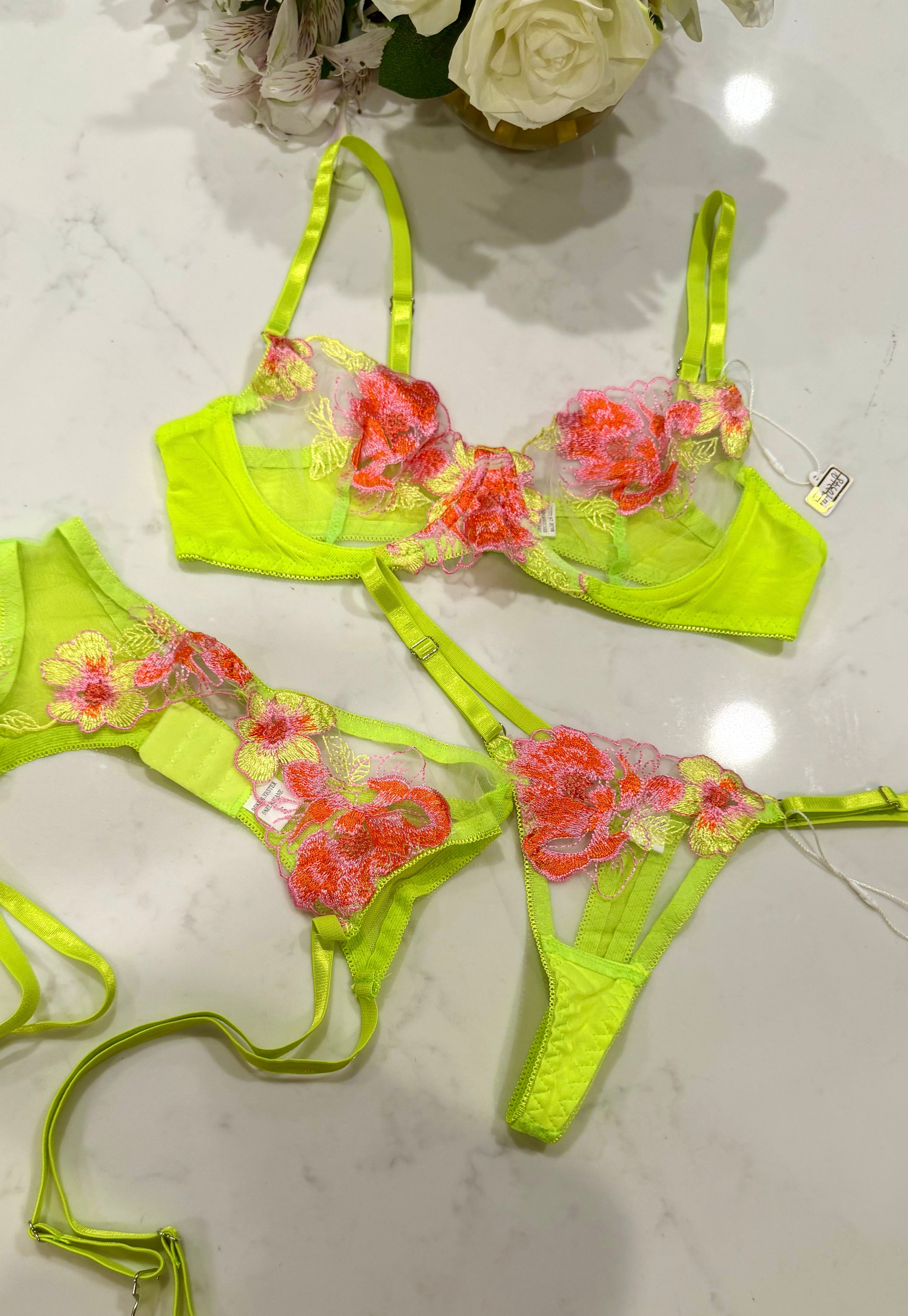 A neon yellow-green lingerie set with floral lace appliques, laid out on a white marble surface. The set includes a bra and matching thong with bright neon yellow-green mesh and straps, decorated with pink, red, and yellow 3D floral lace details.