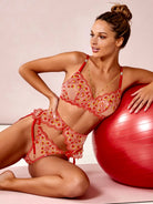 A woman models a red and orange floral lace lingerie set while leaning against a large red exercise ball in a studio setting.