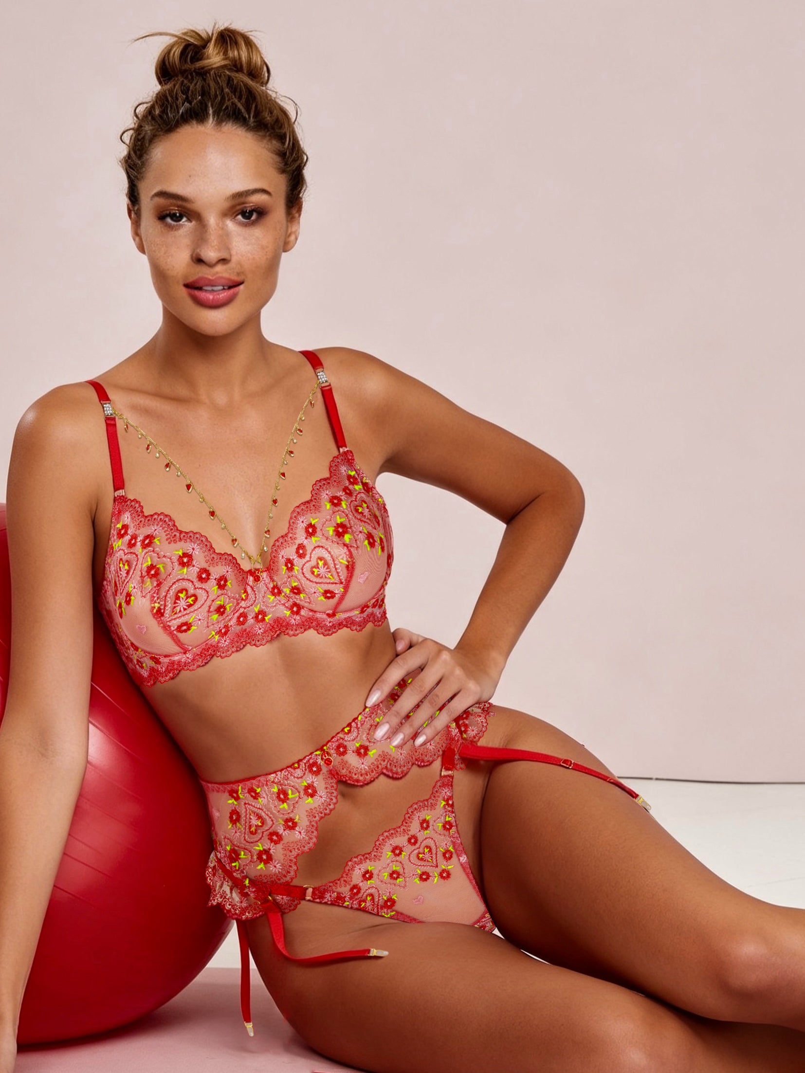 A woman models a two-piece lingerie set consisting of a red and pink paisley-patterned bra and matching g-string bottoms, posed next to a red exercise ball against a light pink background.