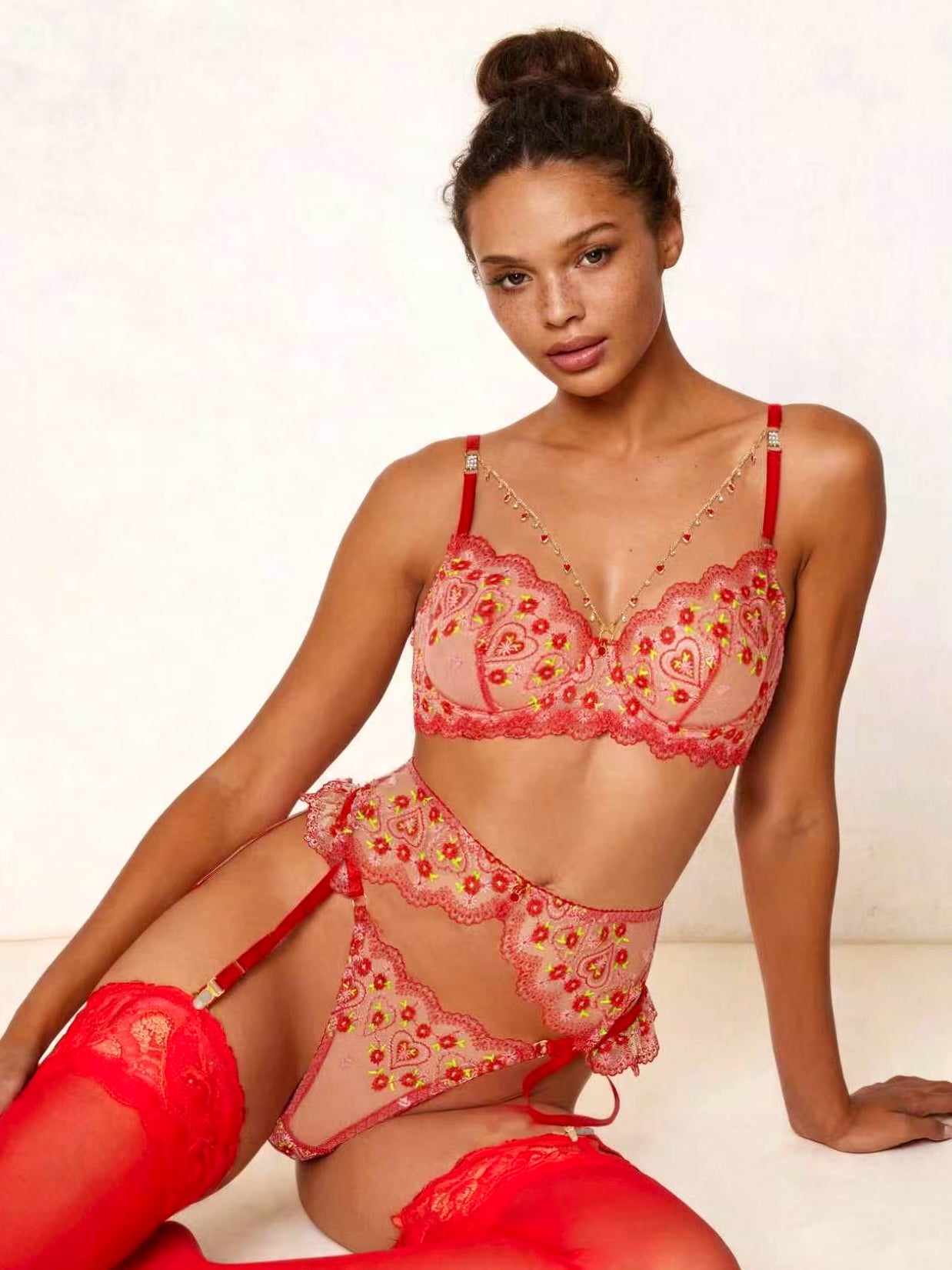 A model wearing a red and pink floral lace lingerie set with matching red thigh-high stockings, posed against a light textured wall.
