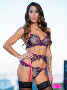 A woman modeling a sheer floral lace lingerie set with bright pink trim and black lace garter details, photographed against a bright outdoor background.