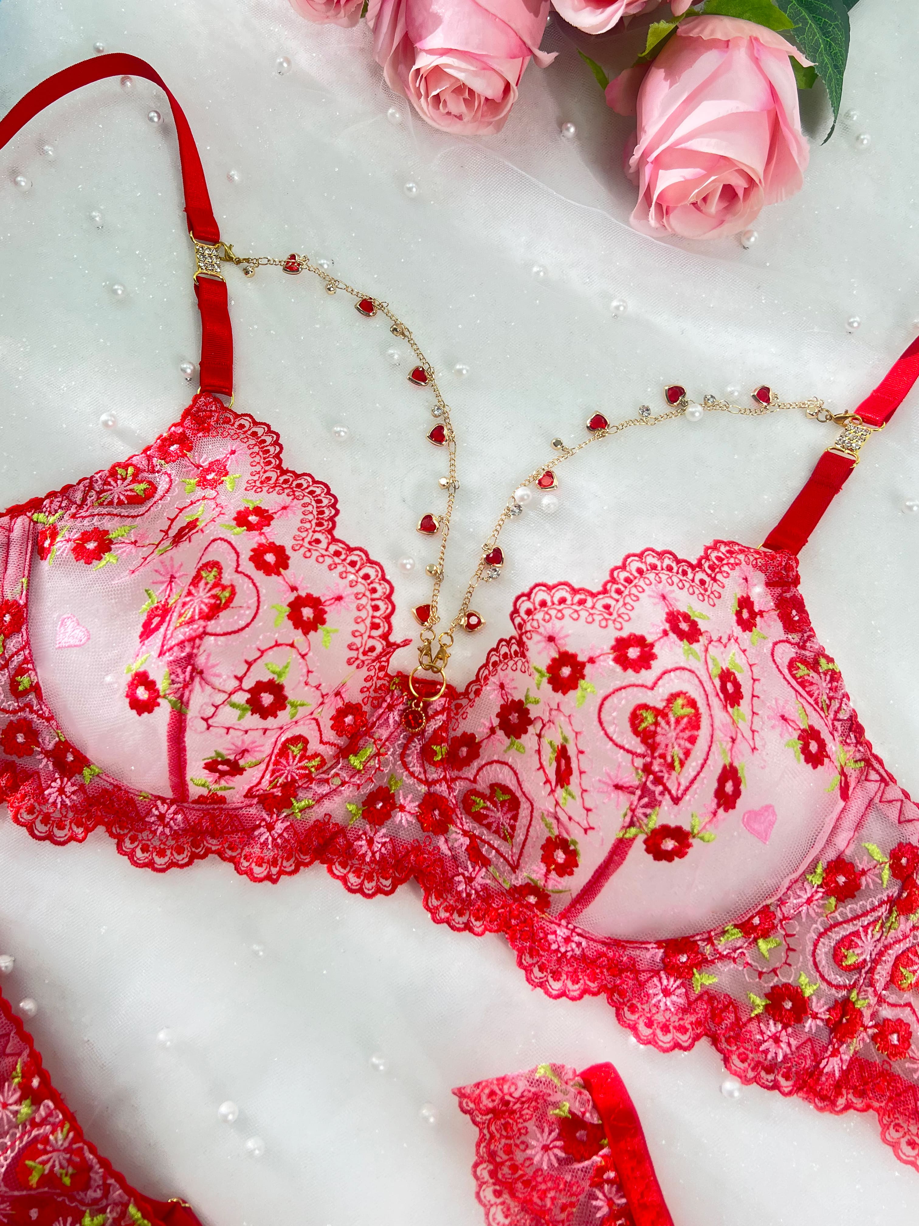 Red and pink lace lingerie set with floral embroidery, heart motifs, and delicate beaded chains, styled with pink roses on a white textured background.