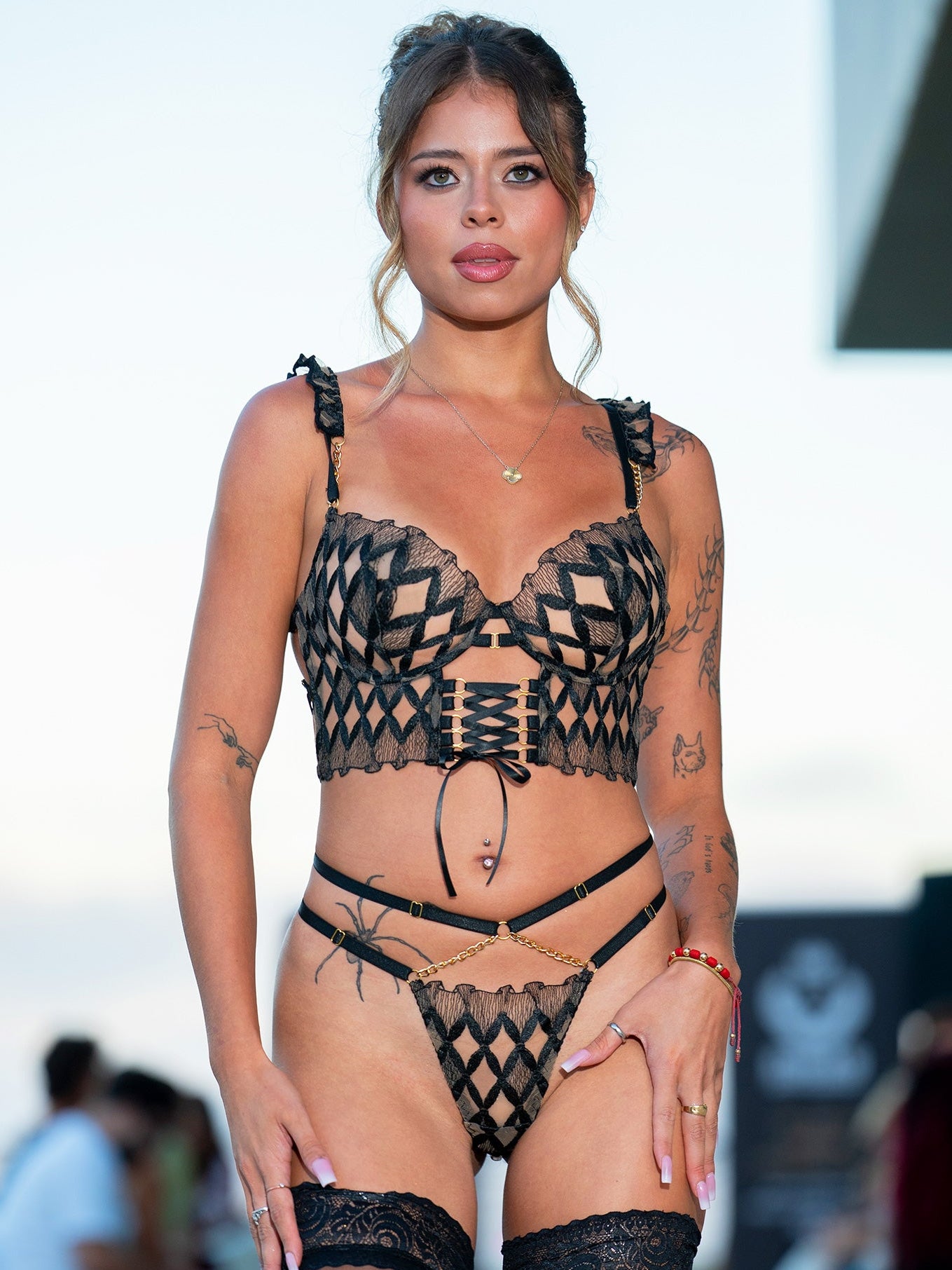 A woman modeling a black lace lingerie set with a geometric pattern, including a bra, thong, garter belt, and thigh-high stockings, set against a blurred outdoor background.
