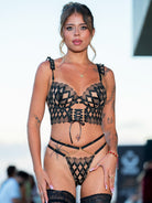 A woman modeling a black lace lingerie set with a geometric pattern, including a bra, thong, garter belt, and thigh-high stockings, set against a blurred outdoor background.