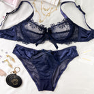 A navy blue lace and satin lingerie set laid flat on a white fabric background, including a bra and matching panties with bow details. Jewelry, a small round black pouch, and a card tag are placed around it as styling props.