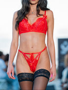 A model walks the runway wearing a bright red lace lingerie set with matching black lace thigh-high stockings against a blurred outdoor background.